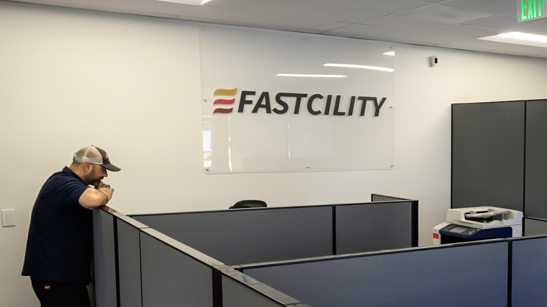 A Fastcility technician leaning on a cubicle partition in the company call centre, with the Fastcility logo mounted on the wall behind.